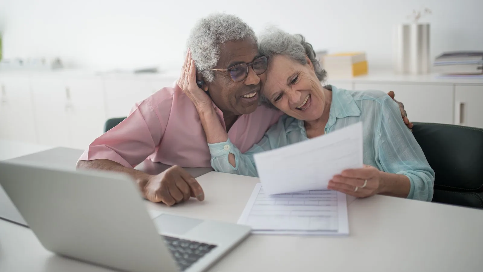 A joyful senior couple smiling while reviewing documents together at a desk with a laptop.