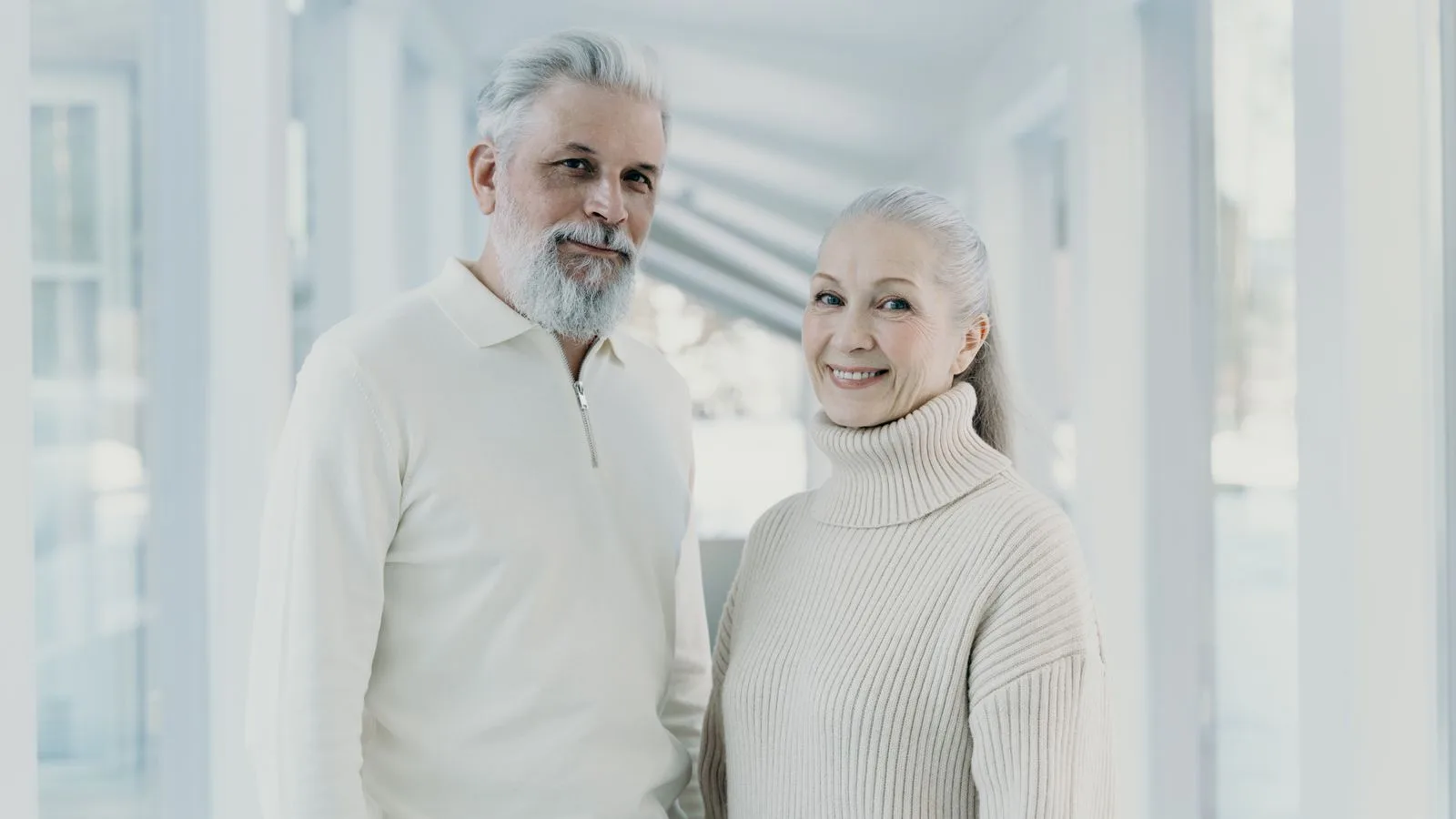 Happy senior couple wearing cozy sweaters, smiling inside a bright, modern space.