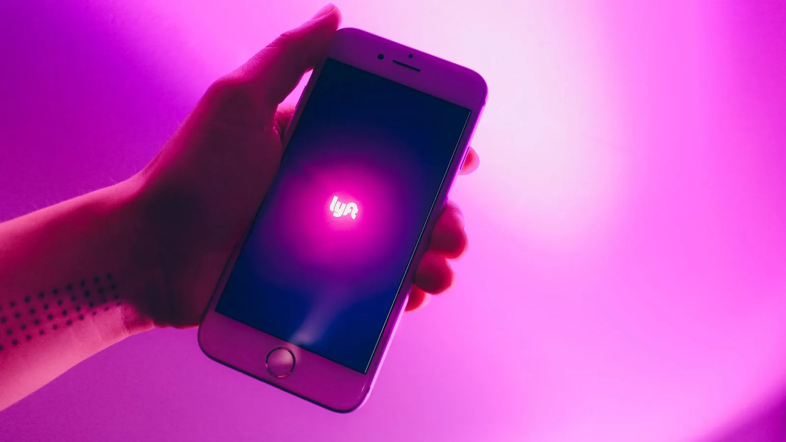 A hand holding a smartphone displaying the Lyft ridesharing app with a vivid pink background.