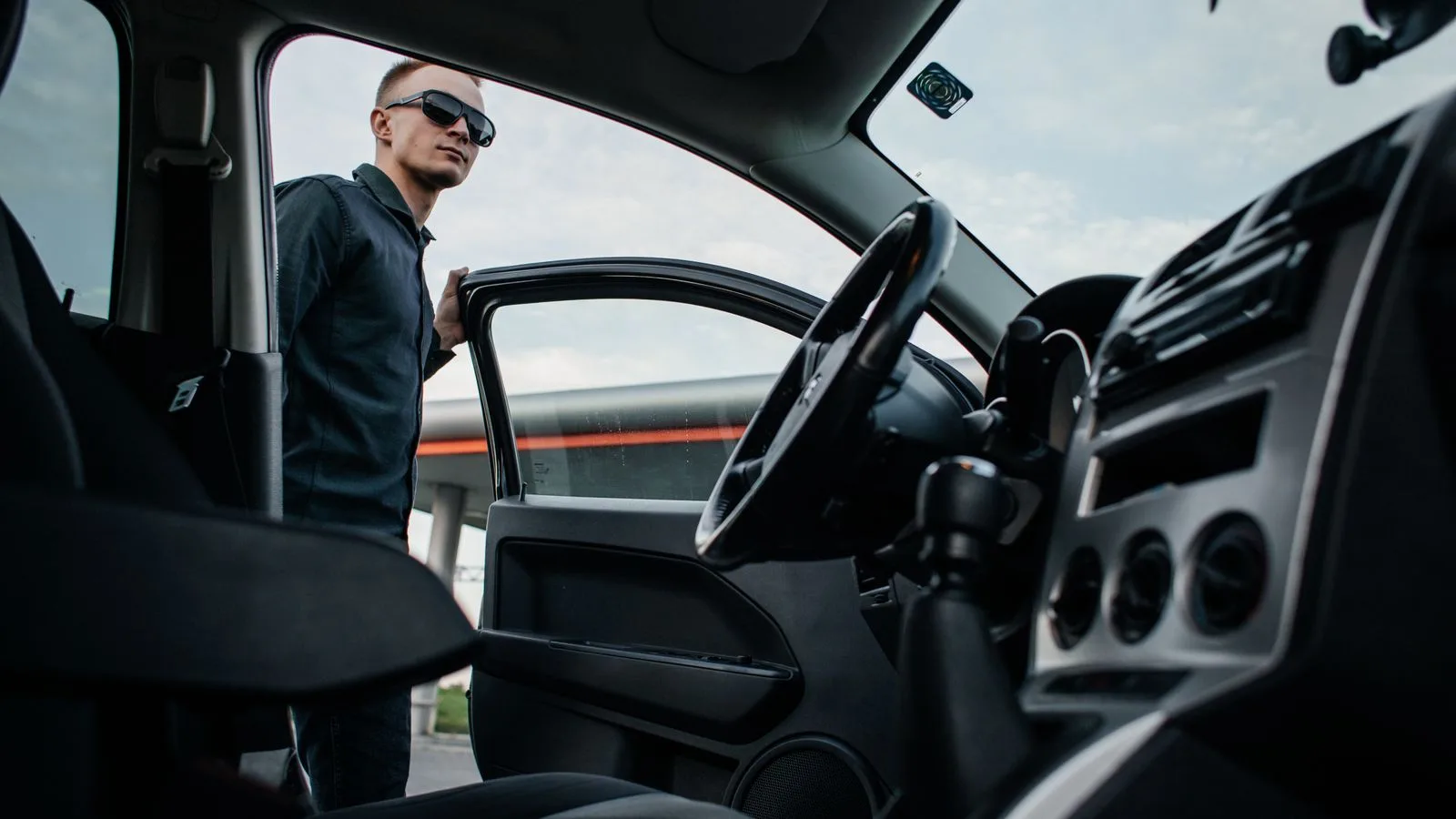 A man in sunglasses stands by an open car door, viewed from the interior.