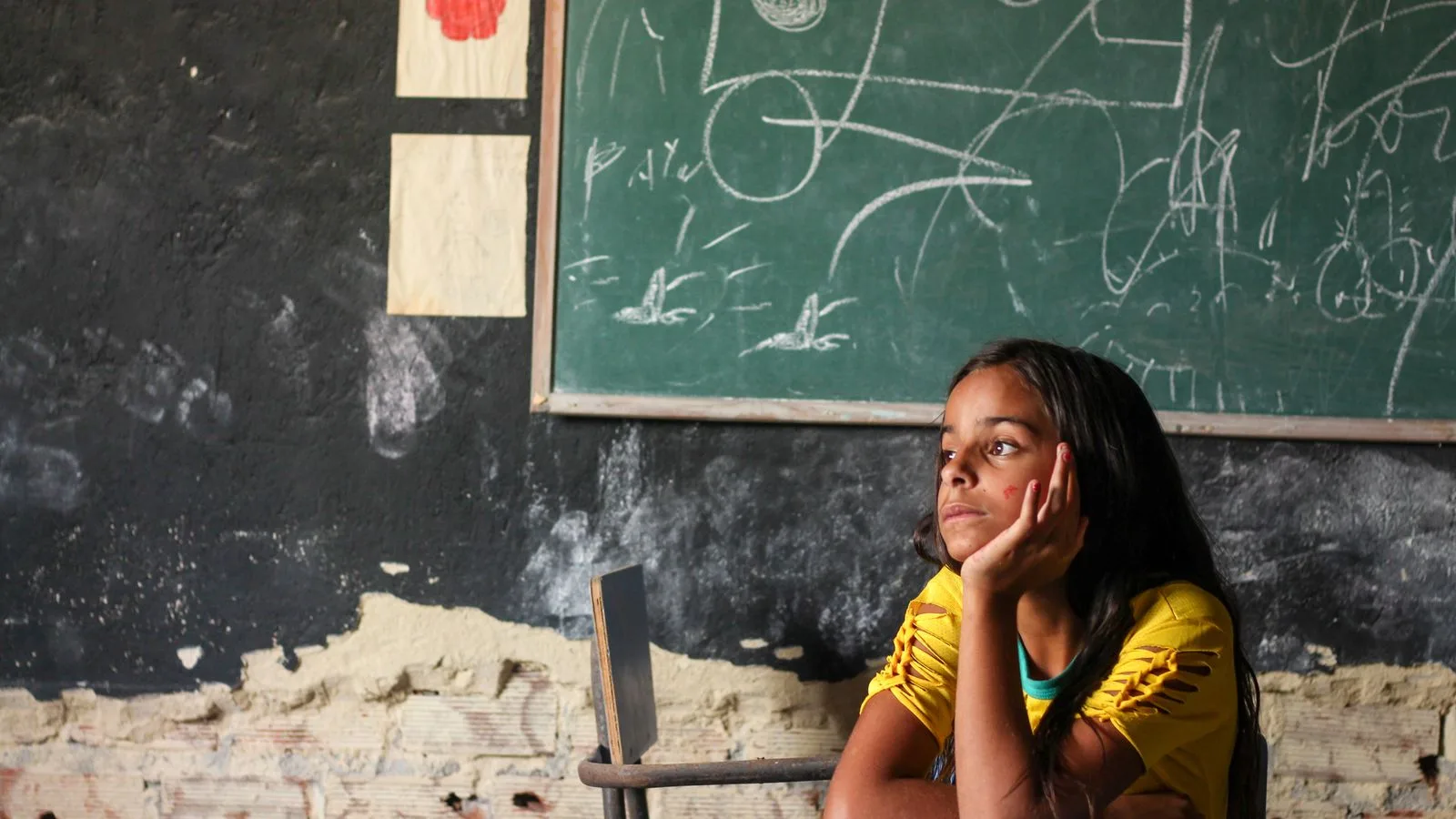 A young girl in a Brazilian classroom sits in thought, surrounded by chalk drawings on a blackboard.