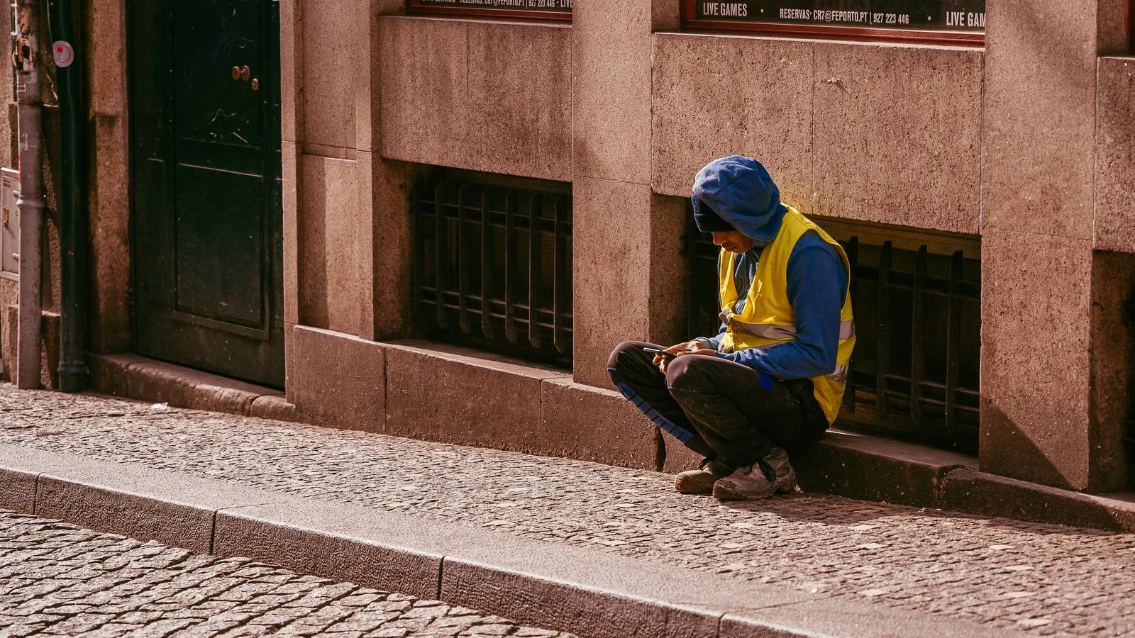 A construction worker in a high-visibility vest sits on a city sidewalk using a smartphone.