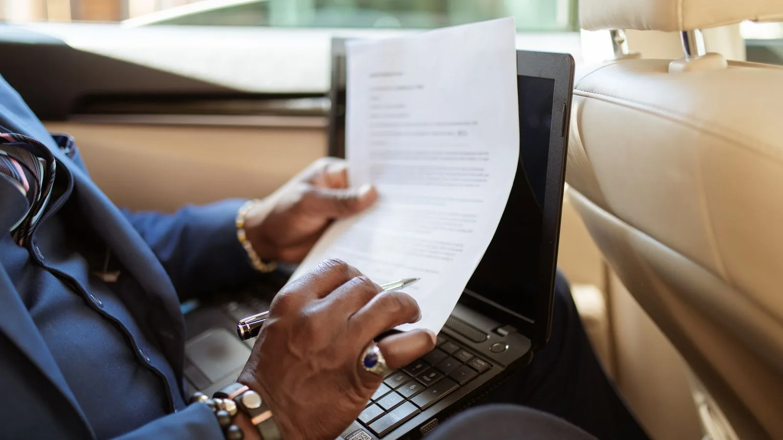 A businessman reviews documents on a laptop while traveling in the backseat of a car.