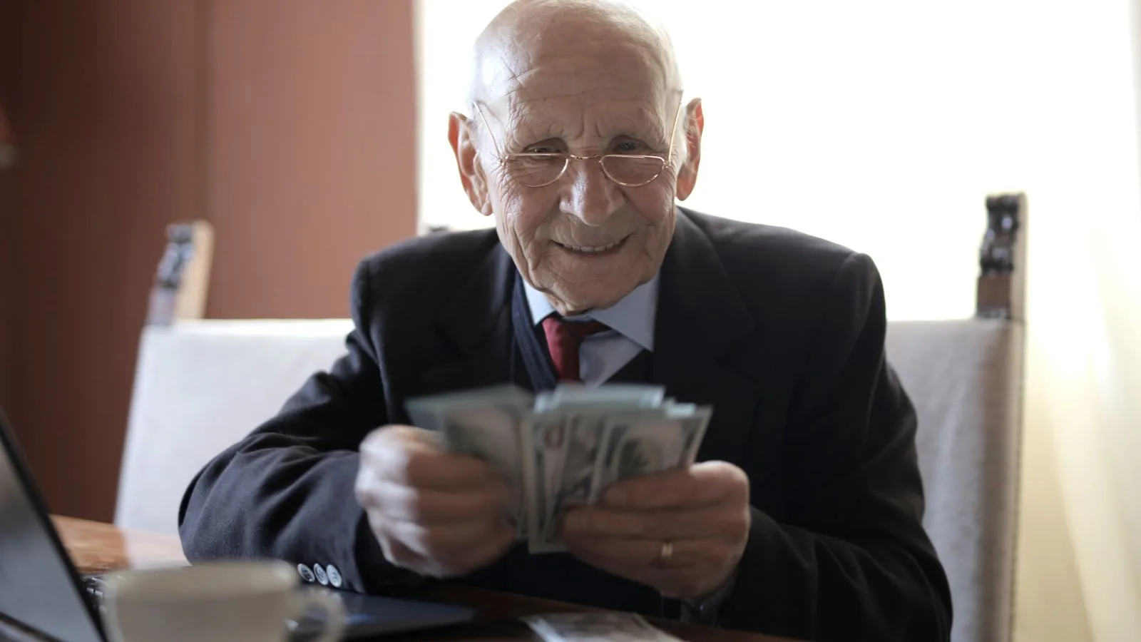 Positive senior businessman in formal suit and eyeglasses counting money bills while sitting at wooden table with cup of beverage and near opened laptop