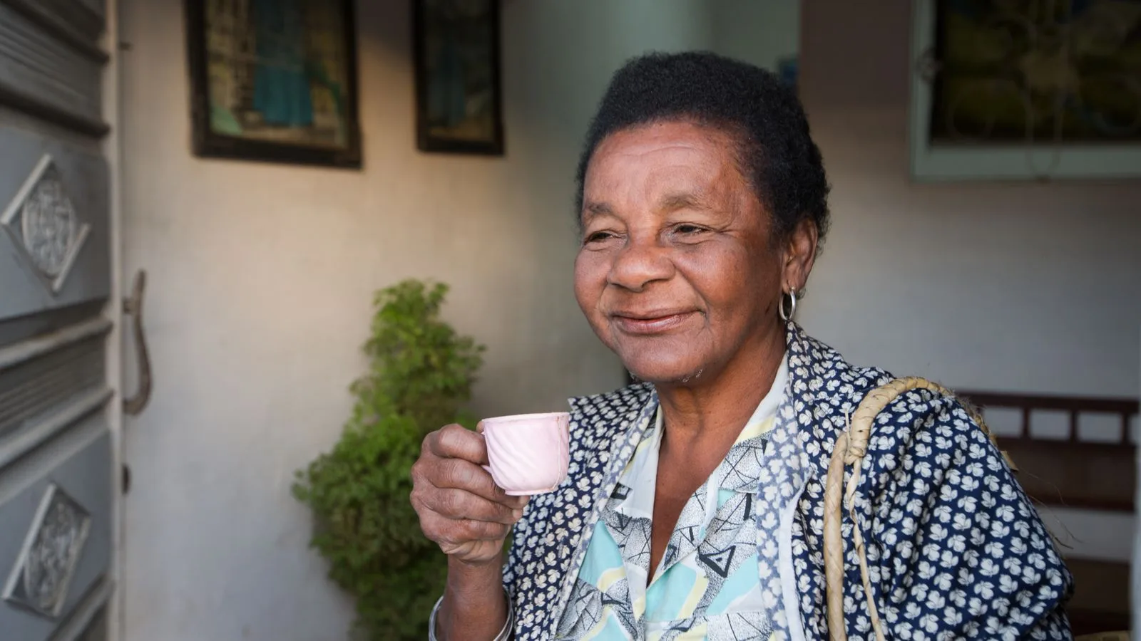 Cheerful ethnic elderly female with short hair in casual clothes standing in room with pictures on walls while drinking coffee in daylight