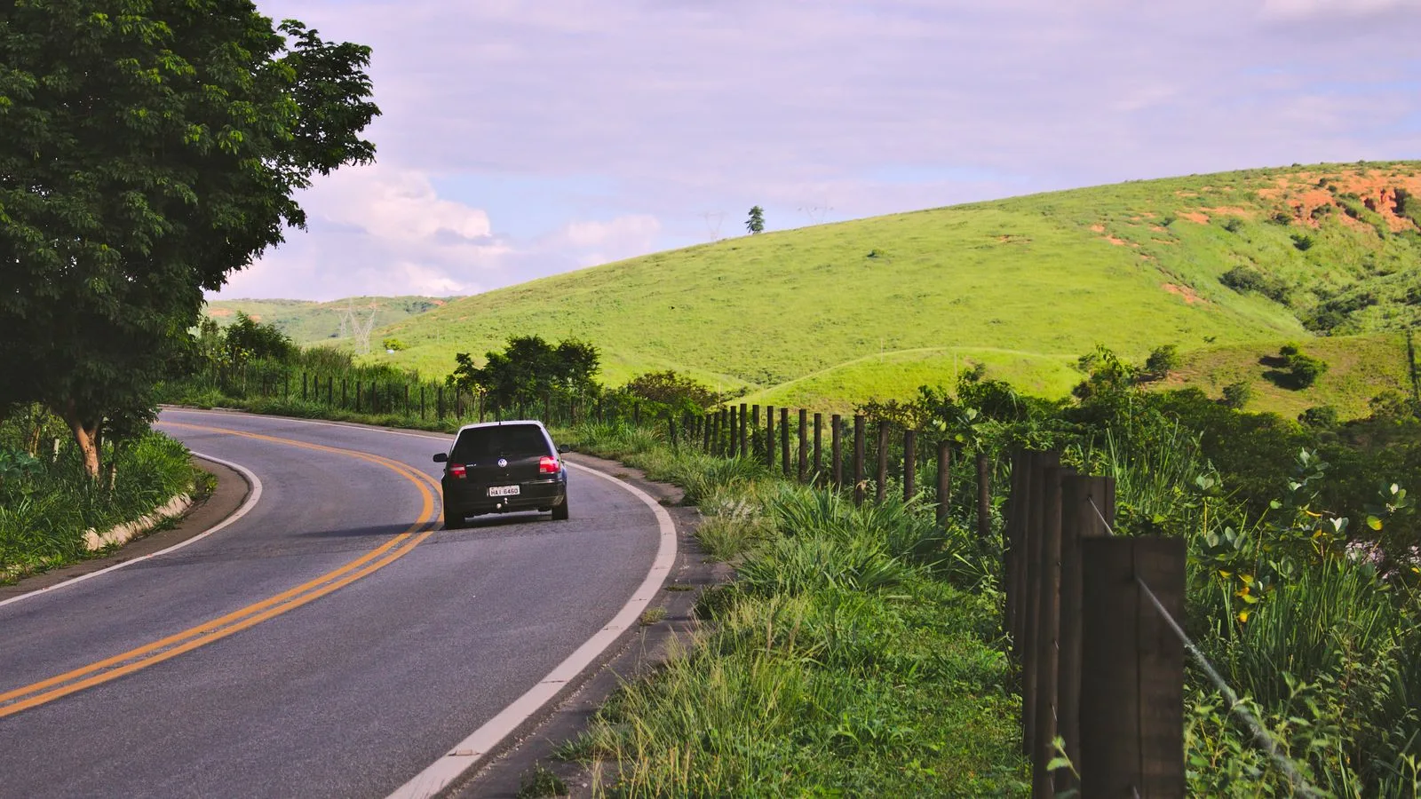 A car travels on a winding rural road in lush Brazilian countryside under a blue sky.