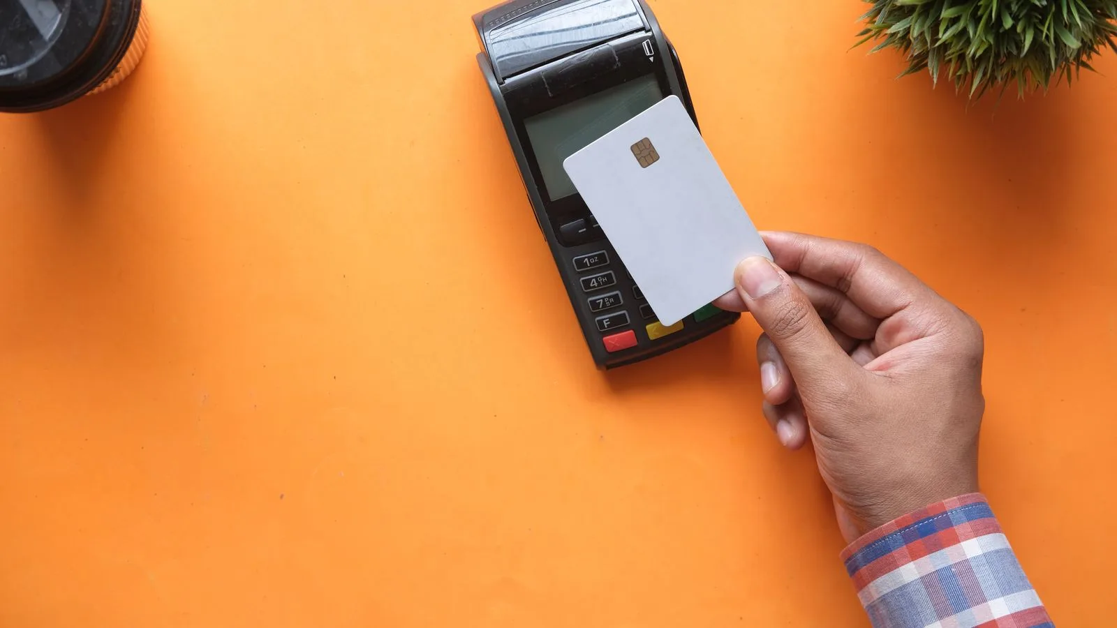 A person making a contactless payment with a credit card and card reader on a bright orange surface.