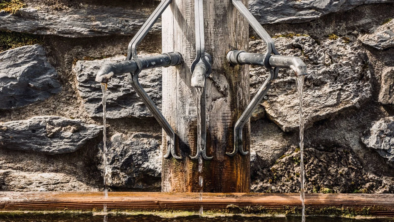 Metal taps of fountain with pouring water into tank placed on wooden post near stone wall on street in city