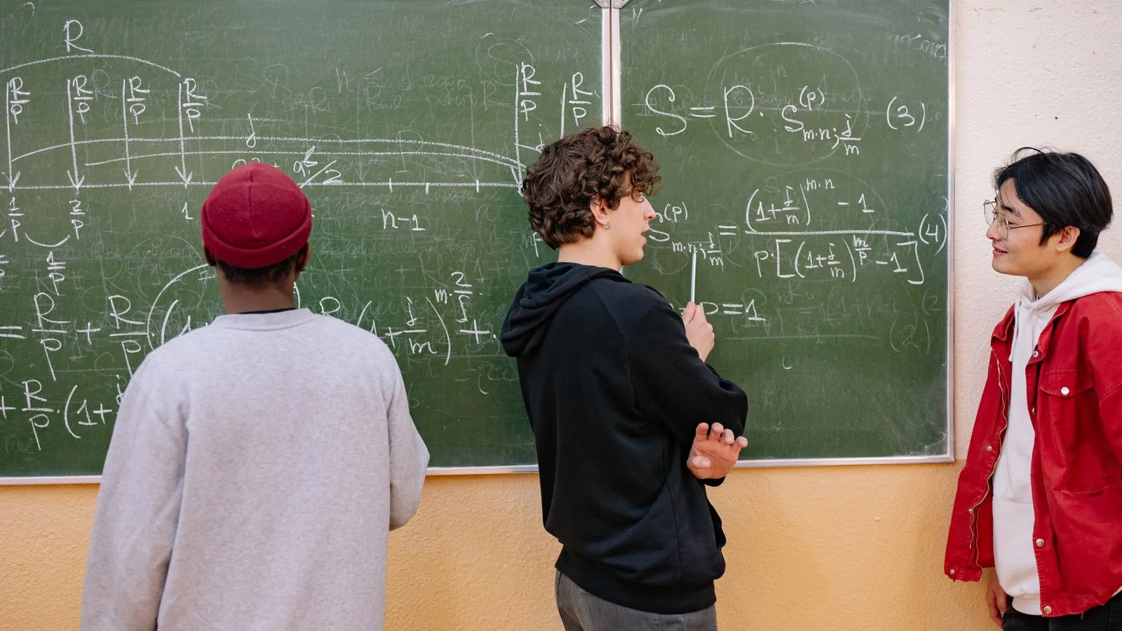 Three students discuss math equations on a classroom blackboard, fostering learning and teamwork.