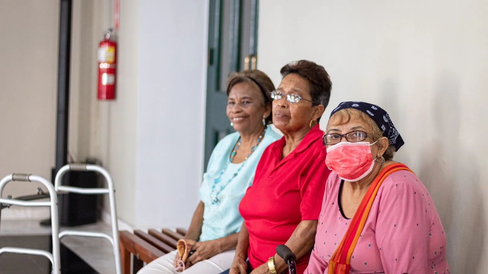 Three senior women sitting indoors, one wearing a face mask, in a safe environment.
