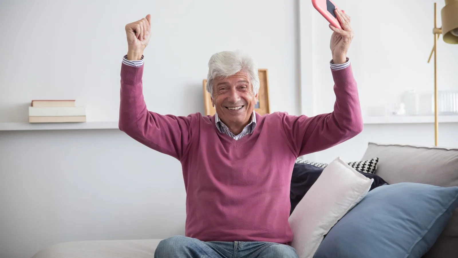A happy senior man sitting on a sofa holding a smartphone indoors, celebrating with raised arms.