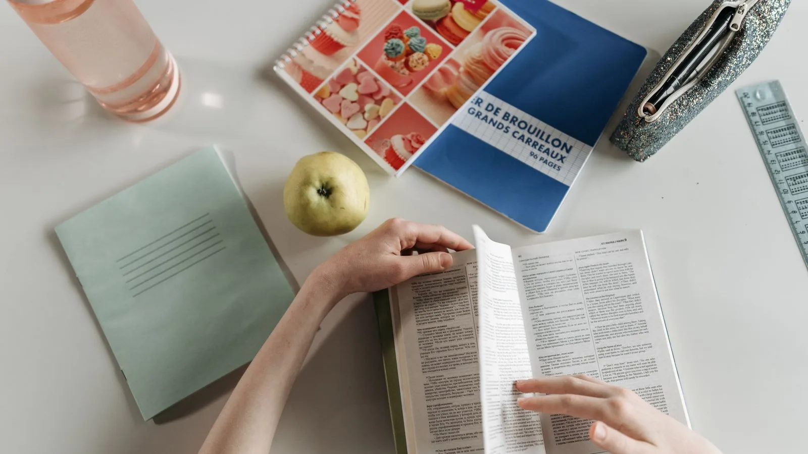 Overhead view of hands turning book pages at a study desk with notebooks, apple, and water bottle.